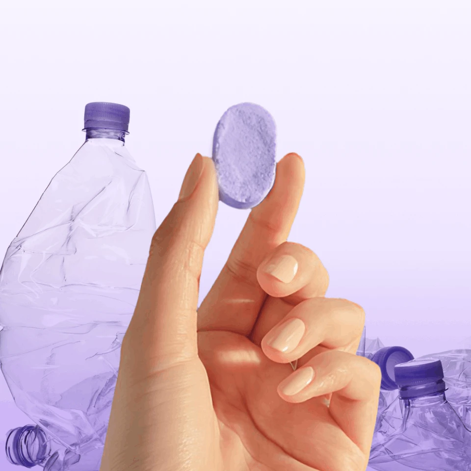 Hand holding a purple cleaning tablet in front of translucent plastic bottles.
